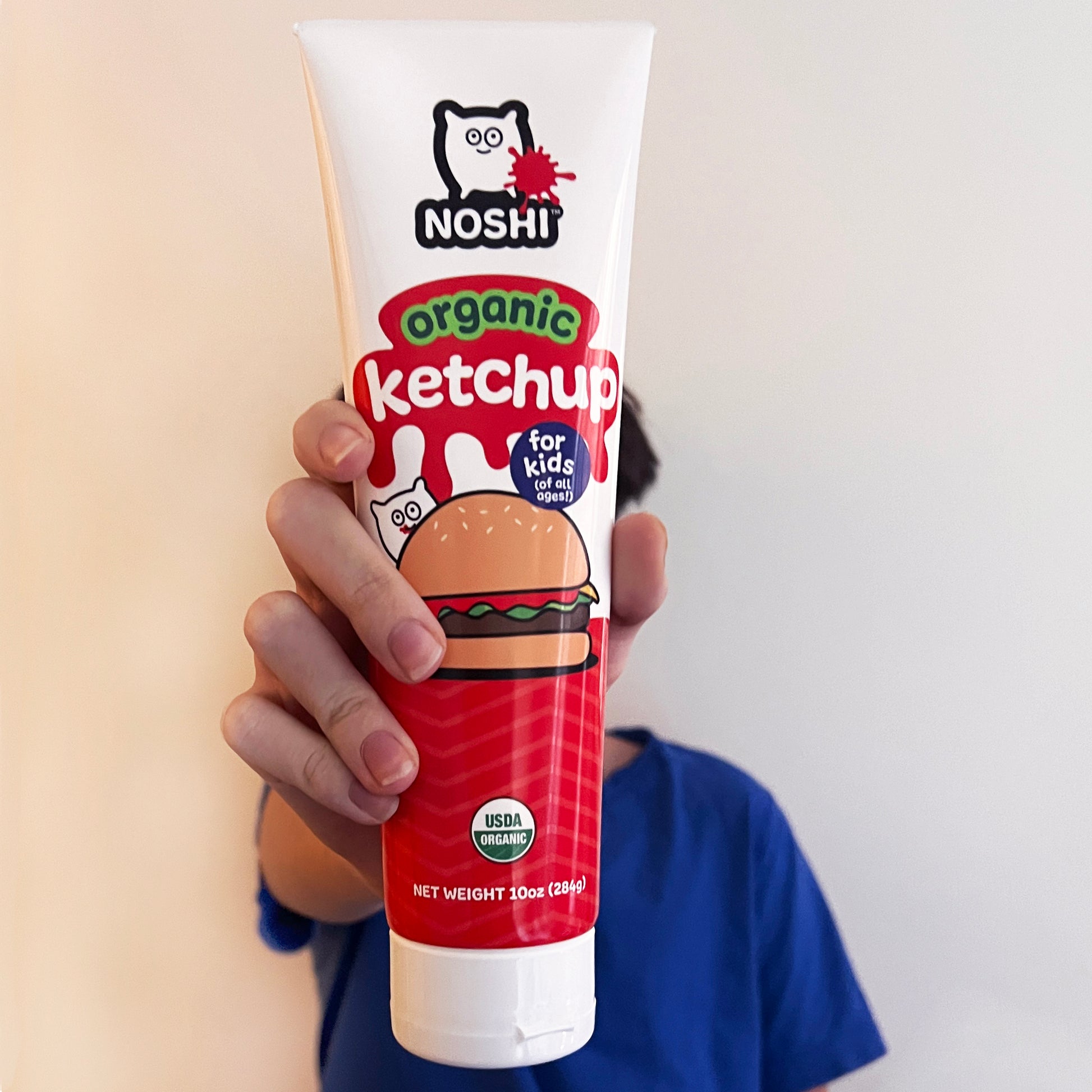 A kid in a blue t-shirt standing in front of a white wall proffers a dispenser of Noshi organic Ketchup for kids - holding it in front  of his face.