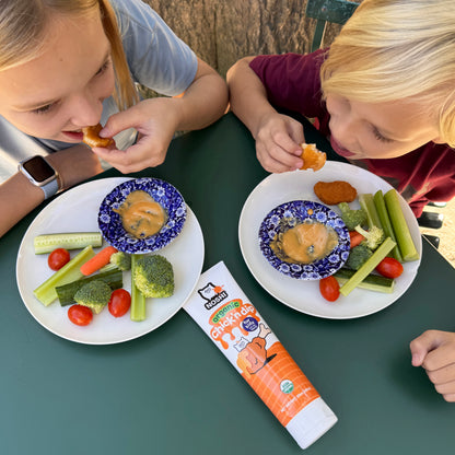 Two kids seen from above sitting at an outdoor table eating chicken tenders and veggies with Noshi organic Chick'n dip, there is a tube of the dip on the table between them