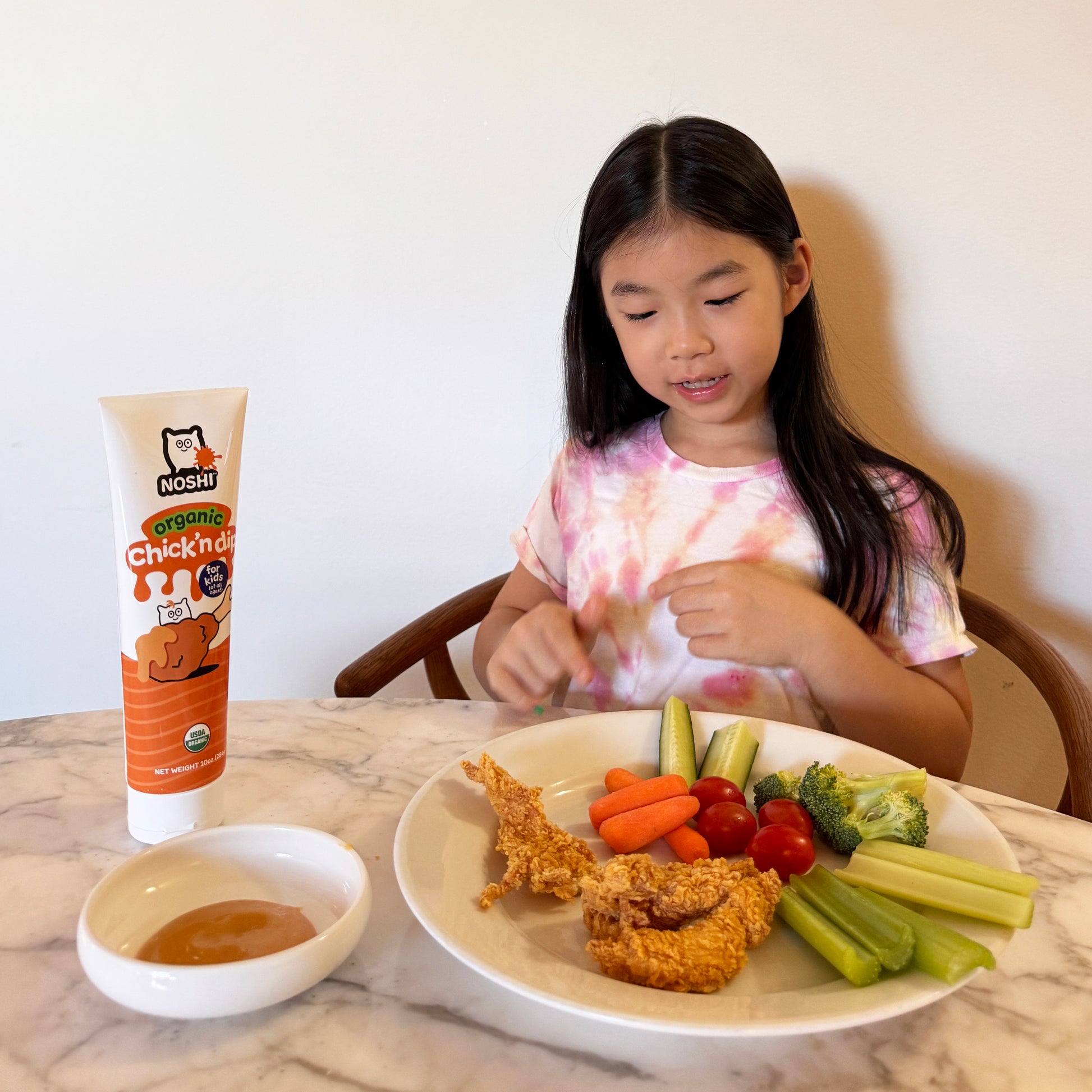 Young girl with long hair and a tie-dye t-shirt looks expectantly at a plate of chicken tenders and veggies with a tube and big bowl of Noshi Chick'n dip on the table beside her.
