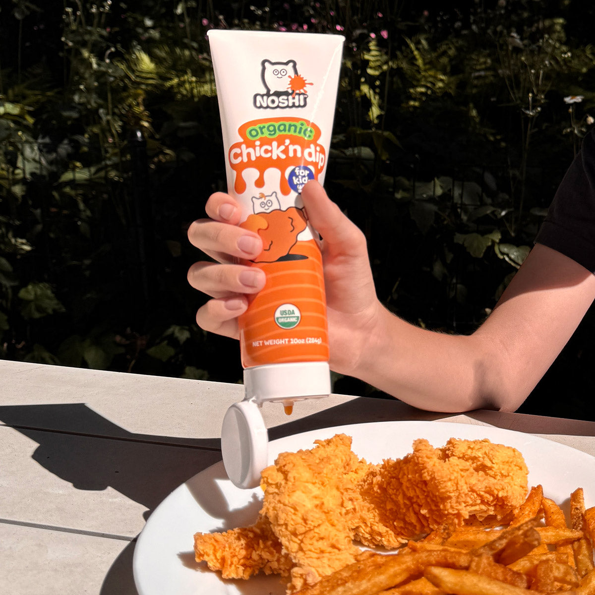 on a sunny day at a picnic table we see a hand holding and squeezing Noshi organic Chick'n dip onto fried chicken and french fries