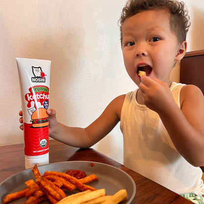 a curly haired toddler eats a ketchup dipped in Noshi organic ketchup with one hand gripping the tube and a plate set before her on a wooden table