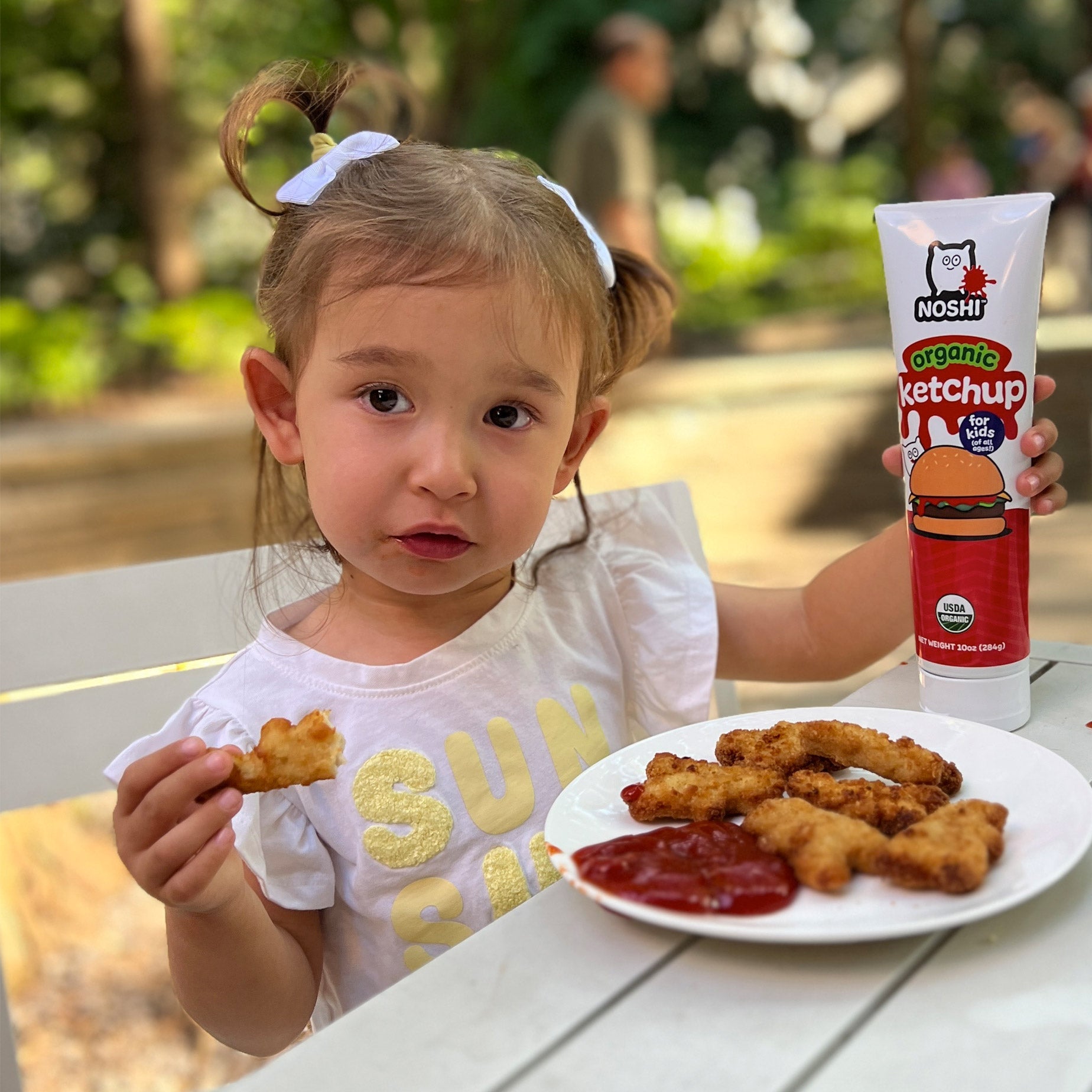 on a sunny day at a picnic table we see a small girl in pig tails  holding a Noshi organic Ketchup which she has just squeezed onto chicken nuggets, she holds a nugget ready to eat in her right hand