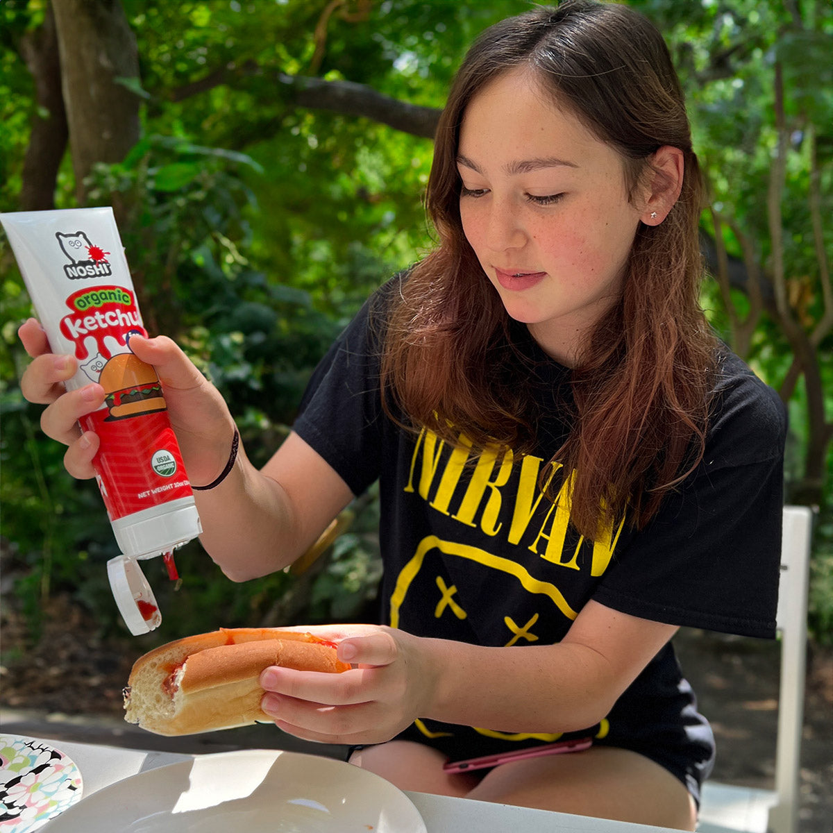 A cool teen girl in a park wearing a  Nirvana t-shirt puts ketchup on a hotdog she is holding in her hand