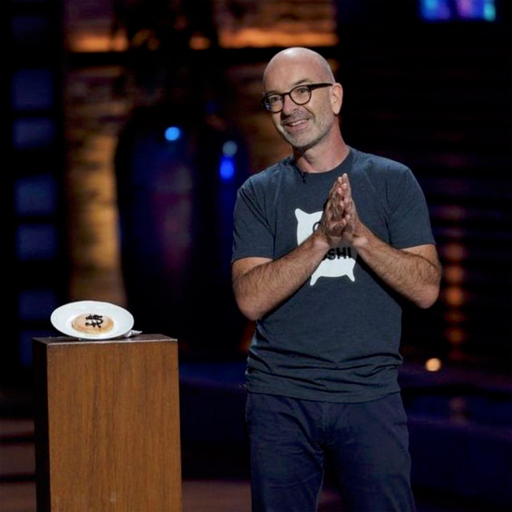 Tomo on stage on Shark Tank next to a plinth with a plate of pancakes painted with a dollar sign in jelly