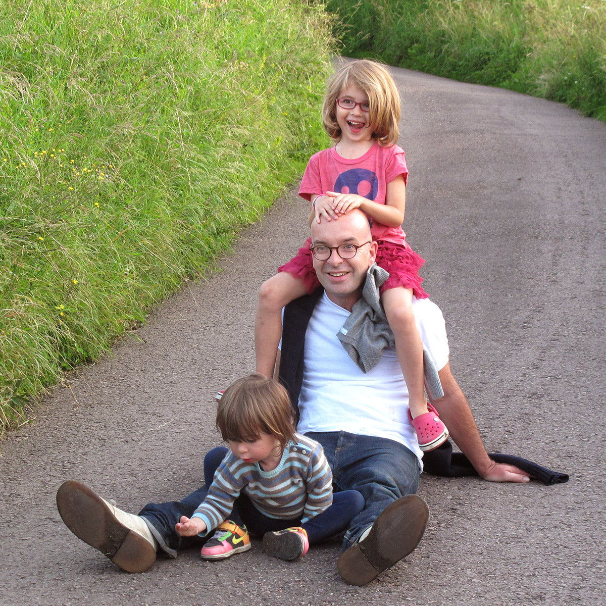 Tomo, the founder of Noshi with his kid. He sits on a gravel road, tall grasses on either side, with one kid on his shoulders smiling with her hands a-top his bald head as a smaller kid sits between his legs examining the gravel