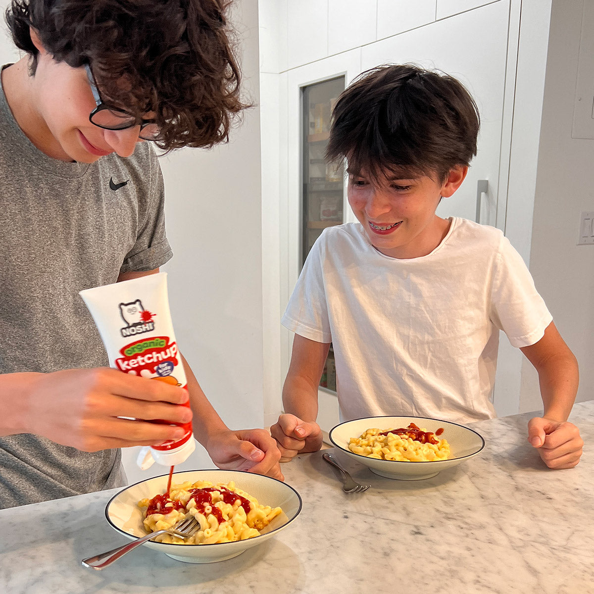 A couple teen/tween boys are at their kitchen counter about to eat mac and cheese with Noshi organic ketchup - the kid on the right has already applied ketchup and the one on the left is mid-squirt